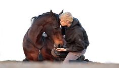 a woman kneeling down next to a brown horse