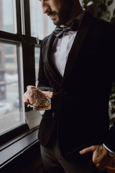 a man in a tuxedo is holding a glass and looking out the window