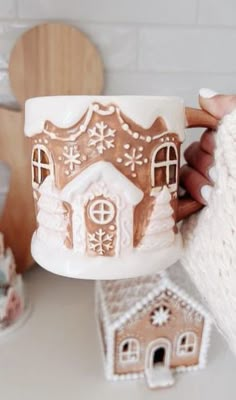 a person holding a cup with a gingerbread house on it and some cookies in the background