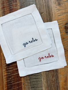 two embroidered napkins sitting on top of a wooden table with the words gorlies spelled in red