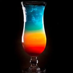 a tall glass filled with colorful liquid on top of a black table next to a dark background