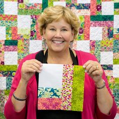 a woman holding up a piece of fabric in front of a quilted wall hanging