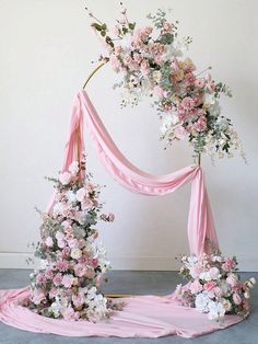 a pink and white wedding arch with flowers on the top, draped over by a light pink fabric