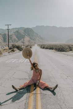 a woman sitting on the side of an empty road with her hat raised in the air