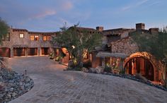 a large stone driveway leading to a house with lots of windows and lights on it