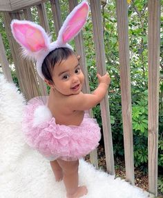 a baby girl wearing a pink tutu and bunny ears, standing in front of a fence