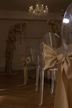 several clear chairs with bows tied around them in a room filled with chandeliers