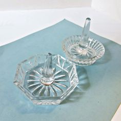 two clear glass dishes sitting on top of a blue tablecloth covered surface, one with a silver lid and the other has a metal spoon in it's center