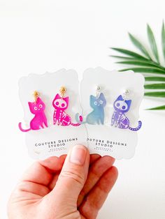 hand holding up three different colored cat earrings on top of a white table next to a plant