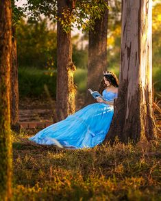 a woman in a blue dress sitting on the ground next to trees and reading a book