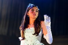 a woman in white dress and gloves on stage with her hand up to the side