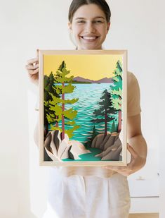 a woman holding up a painting with trees and water in the background, while smiling at the camera