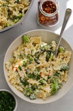 a white bowl filled with pasta and broccoli next to two bowls of seasoning