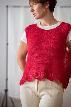 a woman standing in front of a white backdrop wearing a red knitted top and beige pants