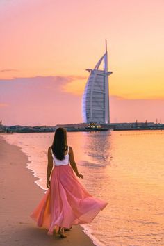 a woman in a pink dress walking on the beach at sunset with burj al arab hotel in the background