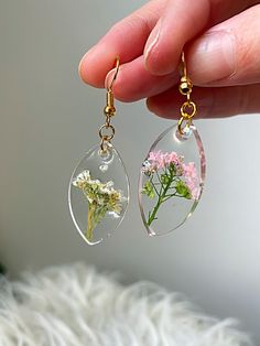 a person is holding two clear glass earrings with flowers inside the dandelion, and one has a tiny flower in it