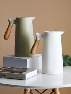 two white vases sitting on top of a table next to a book and plant