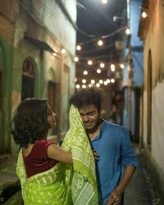 a man and woman standing in an alleyway with lights strung above the buildings behind them