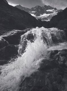 a black and white photo of water rushing over rocks in the mountains with snow on top