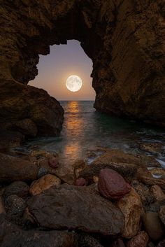 the full moon is seen through an opening in some rocks on the beach at night