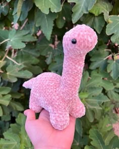 a hand holding a pink knitted toy in front of some green plants and leaves