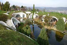 an unusual house on top of a hill next to a pond and forest area with people sitting in it