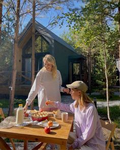 two women sitting at a picnic table with food and drinks in front of a cabin