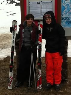 two people standing next to each other holding skis and snowboards in front of a sign