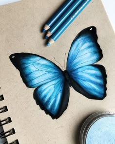 a blue butterfly sitting on top of a notebook next to two pencils and a jar