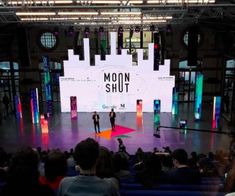 two men standing on stage in front of an audience with the moon shut screen behind them