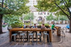 people are sitting at tables in the middle of a city square with trees and benches