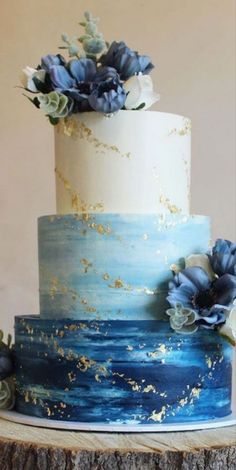 a three tiered blue and white cake with flowers on top is sitting on a tree stump