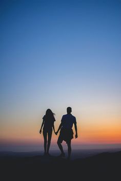 two people are holding hands as the sun sets in the sky behind them on top of a hill