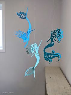 three paper cut mermaids hanging from strings on a wall next to a wooden bench