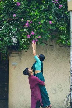 two people reaching up into the air to grab something out of their hands while standing in front of a tree with purple flowers