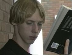 a young man is reading a book in front of a brick wall and looking at the camera