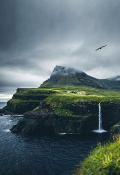 a bird flying over a waterfall on top of a green hill next to the ocean