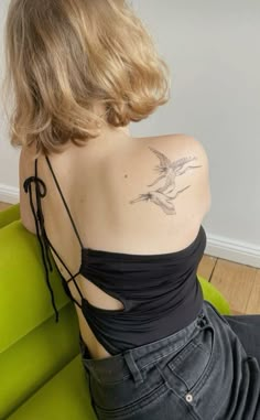 a woman with a bird tattoo on her back sitting on a green couch in front of a white wall