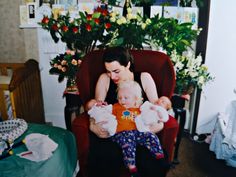 a woman sitting in a red chair holding two babys and looking at the camera