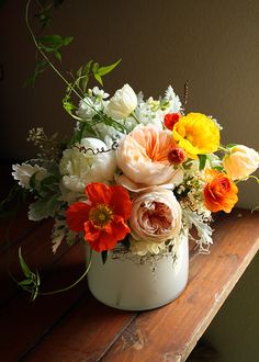 a white vase filled with lots of different flowers on top of a wooden table next to a wall