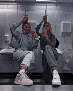 two women taking selfies in a public restroom with their cell phones up to their heads