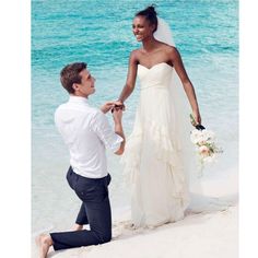 a man kneeling down next to a woman in a wedding dress on the beach holding hands