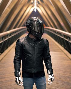 a man wearing a black leather jacket and helmet standing on a wooden bridge with his hands in his pockets