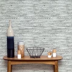 a wooden table topped with two bottles and cups next to a wall covered in woven fabric