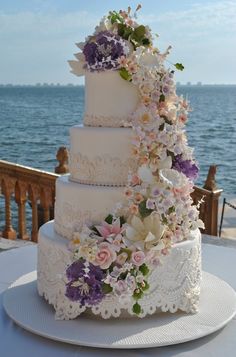 a three tiered wedding cake with purple and white flowers on the top is sitting on a table by the water