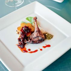 a white square plate topped with meat and veggies next to wine glasses on a table