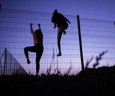 two people jumping up into the air in front of a chain link fence at dusk