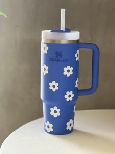 a blue travel mug with white flowers on it sitting on a table next to a plant