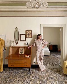 a woman leaning against a wall in a bedroom next to a dresser and bed with clothes on it