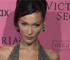 a woman wearing a necklace and earrings on the red carpet at an awards event in front of a pink wall